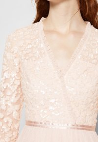 Abito rosa pallido con texture di paillettes, scollatura a V profonda e rifiniture a volant. La cintura in satin e la gonna in tulle stratificato esaltano il design.