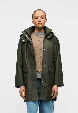 Jeune femme aux tresses cornrows portant un imperméable vert olive sur un cardigan beige et un jean bleu clair, sur un fond uni.