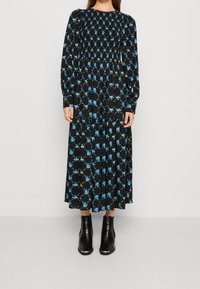 Musta pitkähihaisuus mekko, jossa on sinisen ja vihreän kukkakuosi. Mekossa on smokkayksityiskohta yläosassa ja kerroksellinen hame, yhdistetty mustiin nilkkabuutseihin.