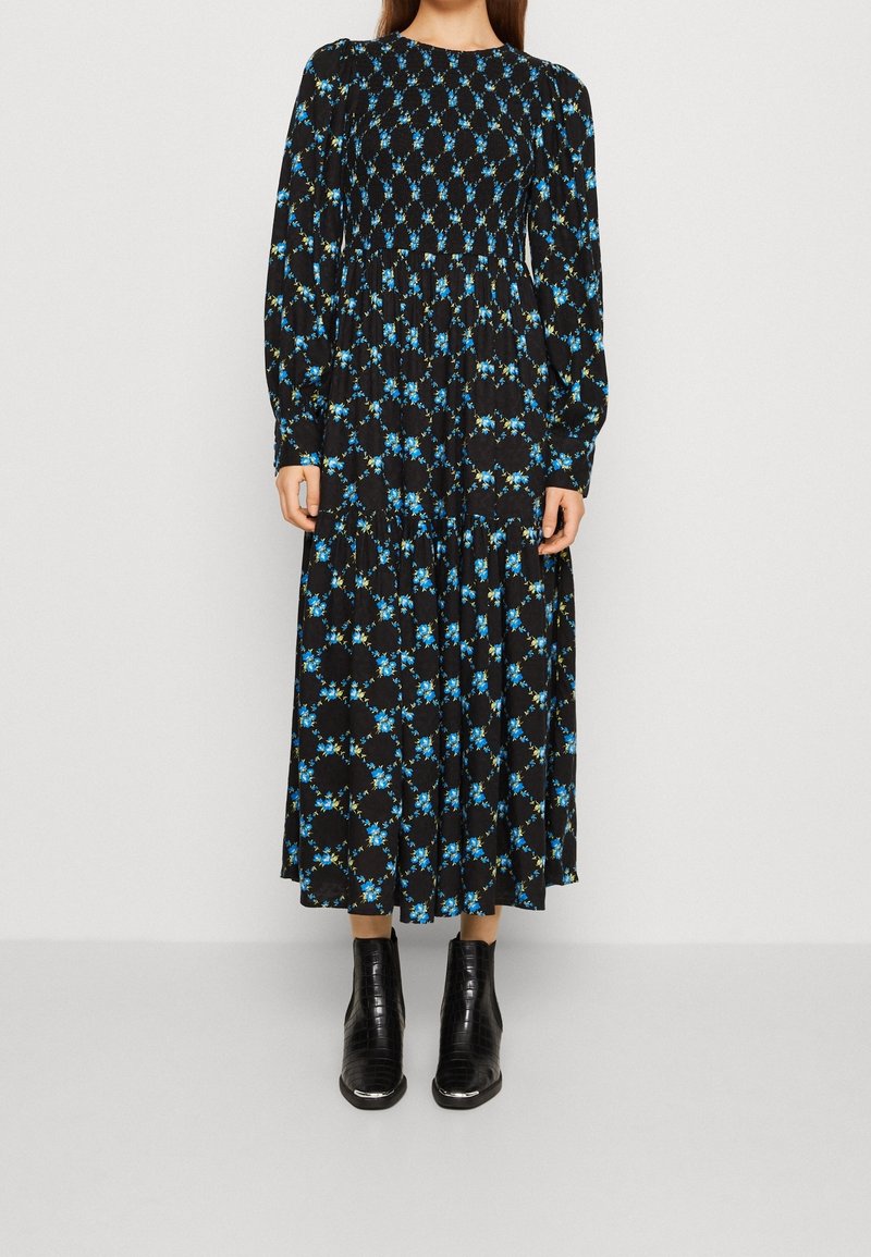 Musta pitkähihaisuus mekko, jossa on sinisen ja vihreän kukkakuosi. Mekossa on smokkayksityiskohta yläosassa ja kerroksellinen hame, yhdistetty mustiin nilkkabuutseihin.