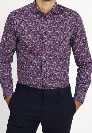Mężczyzna w koszuli z długim rękawem zapinanej na guziki, z czerwonym, niebieskim i białym kwiatowym wzorem, wpuszczonej w ciemnogranatowe spodnie, z rękami w kieszeniach.