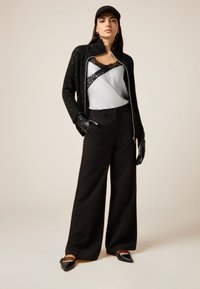 Czarna teksturowana kurtka z srebrnym zamkiem błyskawicznym, nałożona na biały top z koronkowymi wstawkami. Szerokie czarne spodnie i czarne baleriny z detalami.