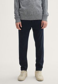 Szürke, textúrázott pulóver bordás mandzsettával, navy slim-fit nadrággal párosítva. A modellen világos bézs sportcipők vannak, semleges háttér előtt áll.