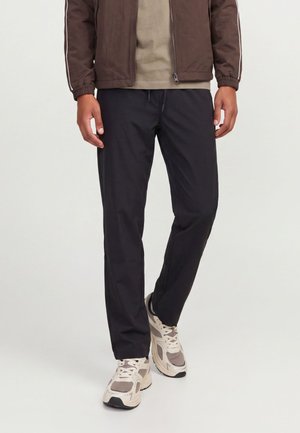 Homme portant un pantalon décontracté noir, des baskets beige et marron, et une veste marron avec des rayures blanches sur les manches, debout sur un fond uni.