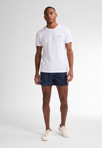 T-shirt bianca in cotone con un piccolo logo rosso, abbinata a pantaloni corti blu navy. Il modello indossa sneakers beige e si trova su uno sfondo grigio chiaro.