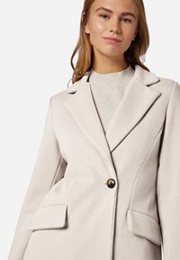 Manteau ajusté beige clair en tissu texturé, avec des revers crantés et une fermeture à un bouton. Comprend deux poches latérales.