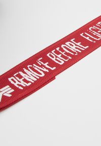 Czerwona wstążka z tkaniny z białym napisem "USUNĄĆ PRZED LOTEM", teksturowana powierzchnia, proste krawędzie oraz mały emblemat z jednej strony.