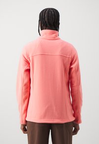 Veste en polaire rose clair avec un col montant, des manches longues et des coutures plates, présentant une texture lisse et un design ajusté.