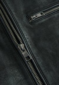 Zwarte leren jas met twee metalen ritsen, waarvan één deels openstaat, waarin een gestructureerd oppervlak en stikdetails zichtbaar zijn.