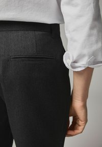Pantaloni din denim închise la culoare, cu o croială ajustată, având o singură buzunar la spate și o textură fină. Purtați cu o cămașă albă cu mânecă lungă.