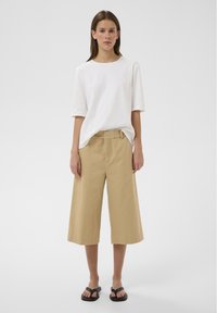 Femme debout portant une chemise blanche à manches courtes, un pantalon beige large et court, et des sandales noires, sur un fond clair uni.