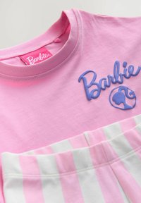 Vaaleanpunainen puuvillapaita, jossa on kohoavaa sinistä "Barbie"-tekstiä ja -kuviota, sekä valkoisia ja vaaleanpunaisia raidoituksia alareunassa.