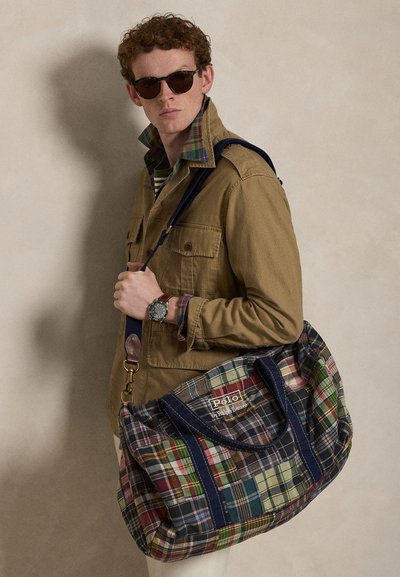 Νέος άνδρας με γυαλιά ηλίου και μπεζ μπουφάν κρατάει μια μεγάλη πολύχρωμη καρό τσάντα Polo στον ώμο, με απλό φόντο.