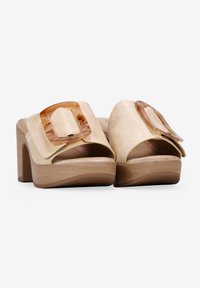 Sandalias de plataforma de ante beige con una gran hebilla translúcida, diseño de punta abierta y tacón de madera. Textura suave y paleta de colores sencilla.