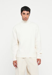 Suéter blanco de cuello alto con un acabado texturizado y puños acanalados. Presenta un sutil detalle de marca en dorado en la manga derecha.