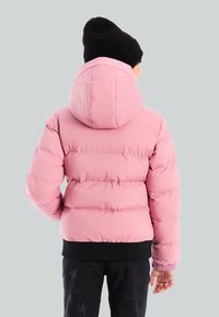 Niño con una chaqueta acolchada rosa con capucha y un gorro negro, de pie con la espalda hacia el espectador sobre un fondo plain.
