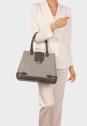 Donna in abito bianco che tiene una borsa beige con motivo, dotata di cinghie e dettagli in pelle marrone scuro, su sfondo chiaro.