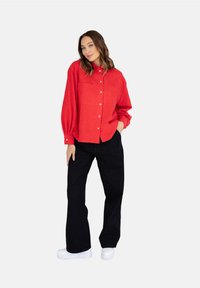 Czerwona koszula zapinana na guziki z dwoma kieszeniami na piersi, długimi rękawami i balonowym krojem, w połączeniu z czarnymi spodniami o szerokich nogawkach i białymi trampkami.