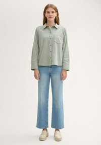 Světle zelená, dlouhá košile s knoflíky a kapsou na hrudi, spárovaná s širokými modrými džíny a béžovými platformovými botami. Jednoduchý a strukturovaný design.