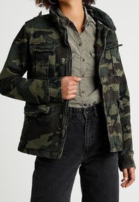 Giacca con motivo camouflage in tonalità verdi, dotata di cappuccio, molteplici tasche e chiusura con zip. Indossata su una camicia grigia chiara con bottoni.
