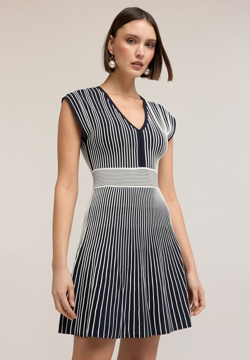 Femeie purtând o rochie bleumarin fără mâneci, cu dungi verticale albe, decolteu în V și fustă plisată, stand pe un fundal bej.