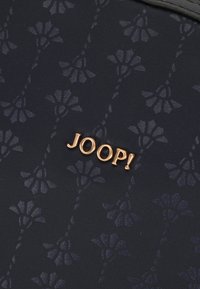 Temno modra tkanina s cvetličnim vzorcem, ki vključuje izrazit zlat "JOOP!" logotip. Tekstura je gladka, z elegantnimi detajli.