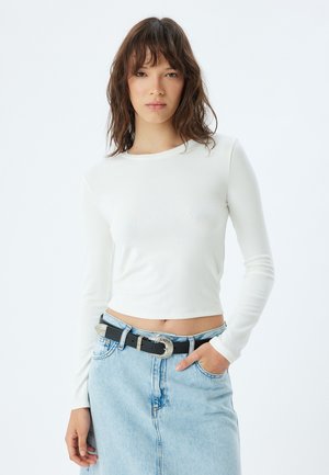 Weißes cropped Longsleeve-Top aus geripptem Material, kombiniert mit einem hellblauen Jeansrock, der einen schwarzen Gürtel mit silberner Schnalle hat.