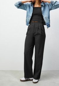 Veste en jean avec poignets côtelés, crop top noir, pantalon noir taille haute plissé et baskets blanches avec accents brun foncé.