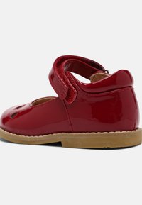 Scarpa in pelle sintetica rossa con punta arrotondata, fascia in velcro regolabile, dettagli intagliati e suola in gomma marrone. Finitura liscia e lucida.