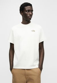 Λευκό βαμβακερό t-shirt με μικρό μπεζ λογότυπο στο στήθος, κοντά μανίκια και στρογγυλή λαιμόκοψη. Λείο ύφασμα, χαλαρή εφαρμογή.