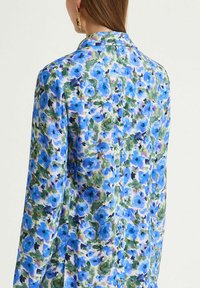 Chemise florale en tissu léger et lisse avec un motif floral bleu agrémenté d'accents verts. Col classique et manches longues.