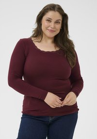 Bordeaux top met lange mouwen en geribbelde textuur, voorzien van een ronde halslijn met kanten afwerking. Gepaard met donkerblauwe jeans, houdt het model de handen op de taille.