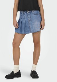 Denim krilo s obrabljenim robom, svetlo modre barve in nabranim dizajnom. V kombinaciji z črnimi loaferji in kremnimi nogavicami.