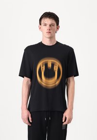 Sort bomulds T-shirt med et sløret gult smilende ansigt i midten. Korte ærmer og en afslappet pasform.