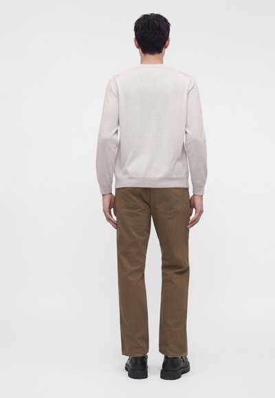 Homme portant une chemise beige clair à manches longues, un pantalon marron et des chaussures noires, debout de dos sur un fond gris uni.