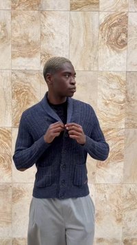 Mladý muž s krátkými blond vlasy se zapíná do tmavě modrého pleteného svetru, má na sobě světle šedé kalhoty a stojí proti béžové mramorové zdi.