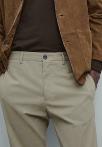 Man i beigefärgade byxor, brun mockajacka och mörkbrun skjorta med händerna i fickorna mot en neutral bakgrund.