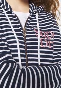 Felpa con zip a righe blu navy e bianche, con un cappuccio e lacci bianchi. Presenta il logo "Superdry" ricamato in rosa e un dettaglio della zip dorata.