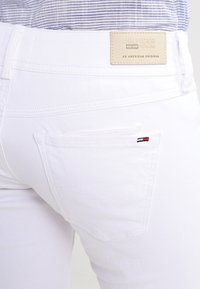Pantalones vaqueros de mezclilla blanca con un diseño cosido y un parche de cuero marrón con marca. Presenta un pequeño logo de color en el bolsillo trasero.