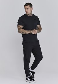 Schwarzes Rundhals-T-Shirt und enganliegende schwarze Hose, kombiniert mit schwarz-weißen Turnschuhen. Die Haut ist tätowiert, vor neutralem Hintergrund.