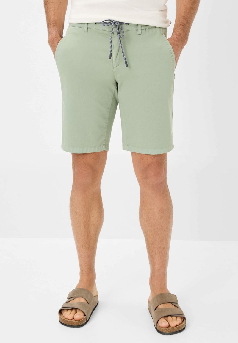 Pantaloni corti verde chiaro con vita regolabile, tasche laterali e una texture liscia, abbinati a sandali scivolati marroni.
