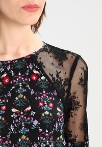 Robe noire à motifs floraux avec des manches en dentelle délicate. Présente un col rond et des motifs vibrants de fleurs et d'oiseaux en plusieurs couleurs.
