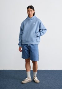 Lyseblue hættetrøje i blødt materiale med en lomme foran og et logo. Båret sammen med denimshorts og sneakers i neutrale nuancer.