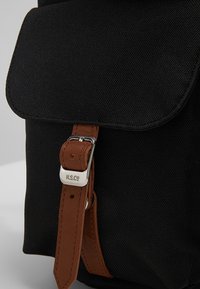 Zwarte stoffen rugzak met een afgeronde klep, leren riem met een zilveren gesp en een reliëflogo. Getextureerd materiaal met een stevig ontwerp.