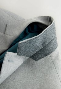 Blazer gris claro con un patrón texturizado, cuello de fieltro gris oscuro y forro interior con diseño en verde azulado, exhibido sobre una superficie clara.