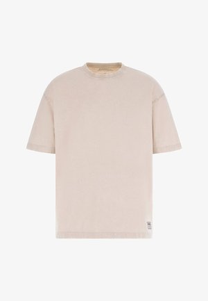T-shirt beige à manches courtes et col rond avec un petit écusson rectangulaire près de l'ourlet inférieur sur fond blanc.