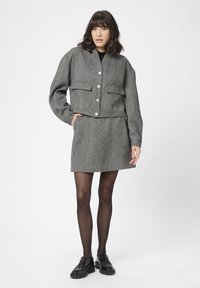 Grijze houndstooth cropped jas met knoopdetails en twee voorzakken, gecombineerd met een bijpassende rok. Het model draagt zwarte maillots en schoenen.