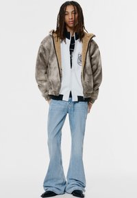Veste bomber en fausse fourrure marron sur un hoodie zippé blanc, associée à un jeans évasé bleu clair et des chaussures noires, se tenant devant un fond blanc.