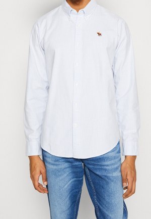 Mann trägt hellblaues, vertikal gestreiftes Hemd mit kleinem braunem Tier-Logo auf der Brust, kombiniert mit blauen Jeans.
