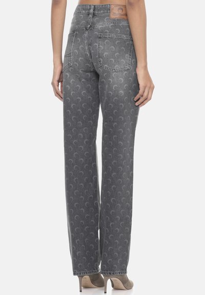 Veduta posteriore snella di una persona che indossa jeans grigi a gamba dritta con motivo e scarpe con tacco taupe, in piedi su uno sfondo bianco.